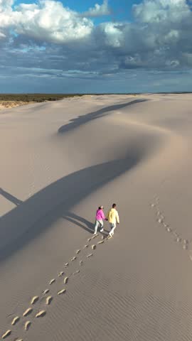 Two people walk hand in hand along the expansive sandy dunes of Rabjerg Mile Jutland, Denmark during golden hour. The soft shadows and warm light create a peaceful