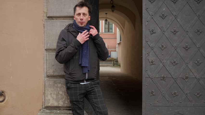 A man posing for a portrait outdoors on a chilly winter day tries to stay warm. He looks at the camera while adjusting his cozy scarf and putting his hands in his pockets for warmth.