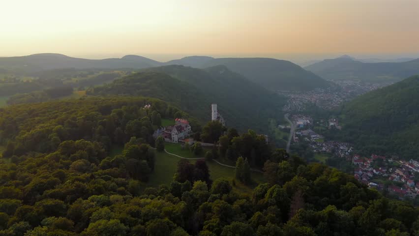 Aerial sunset footage of Lichtenstein Castle, perched on the Albtrauf ridge above Honau, Germany. This romantic mountain fortress glows in warm light, surrounded by forest and steep cliffs.