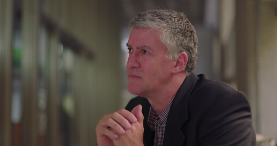 Middle aged man looking into distance with anguished expression, seated in office lounge with hands clasped in front of face in moment of inner turmoil