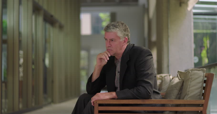 Middle aged man sitting quietly in office lounge with hand on chin, appearing thoughtful and reflective in peaceful modern workspace