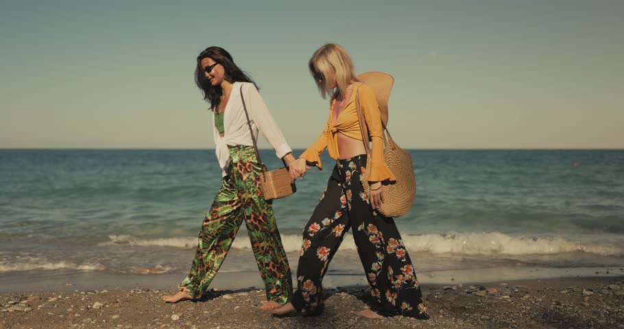 Two stylish women in colorful outfits walk barefoot along a sunlit beach, holding hands and laughing. Ideal lifestyle and travel footage for friendship, summer fashion, or promo content.