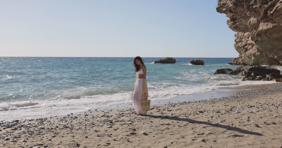 Trendy young woman in beige skirt and white top strolls barefoot along a scenic pebble beach in super slow motion. Ideal for beauty, fashion, travel or lifestyle promo and ads.