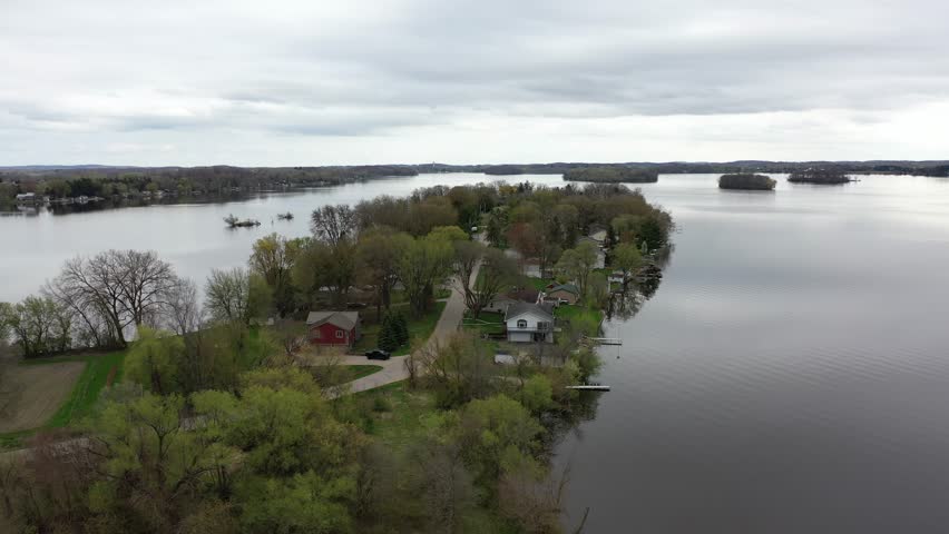 Aerial view of a quiet lakeside neighborhood on a cloudy spring day in rural Wisconsin, showcasing homes, winding roads, bare trees, and calm water under an overcast sky.