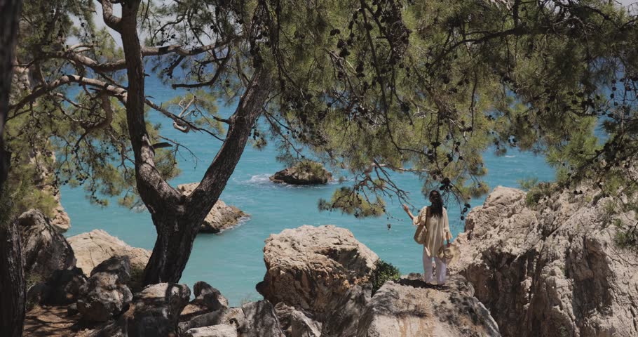 A serene woman stands quietly on rocky cliffs framed by pine trees, gazing at the vibrant turquoise ocean. Stunning wide-angle shot ideal for travel, nature, lifestyle, or wellness content.