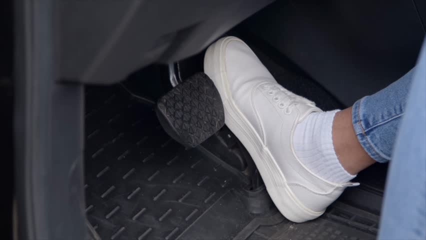 Close-up of female legs in stylish sneakers pressing car brake pedal, while woman driver driving vehicle in city traffic.