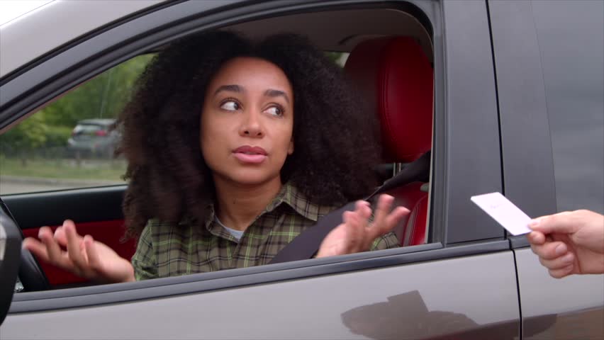 Beautiful African American female driver with Afro hairstyle handing driver's license to police officer during traffic control on road, while woman driving car on trip.