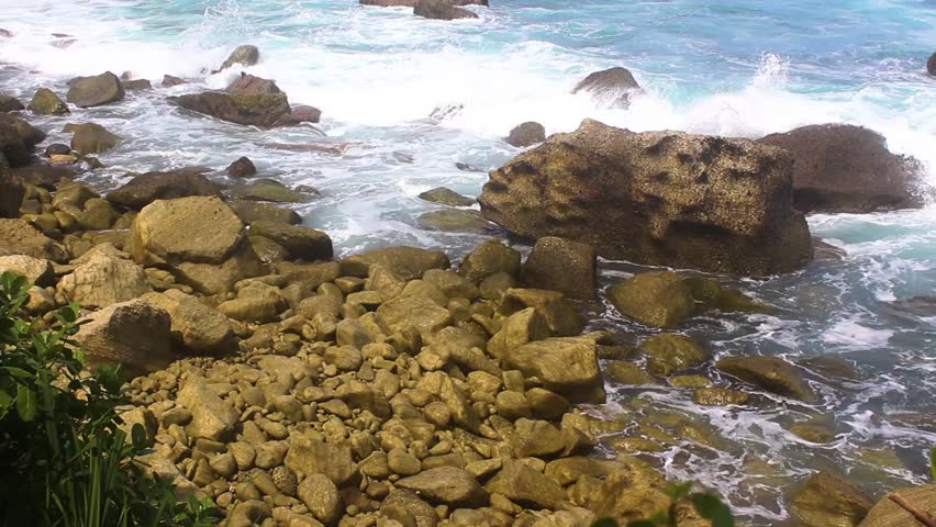 Waves crashing onto a rocky tropical shoreline with large coral-covered boulders and green plants in the foreground under bright daylight.