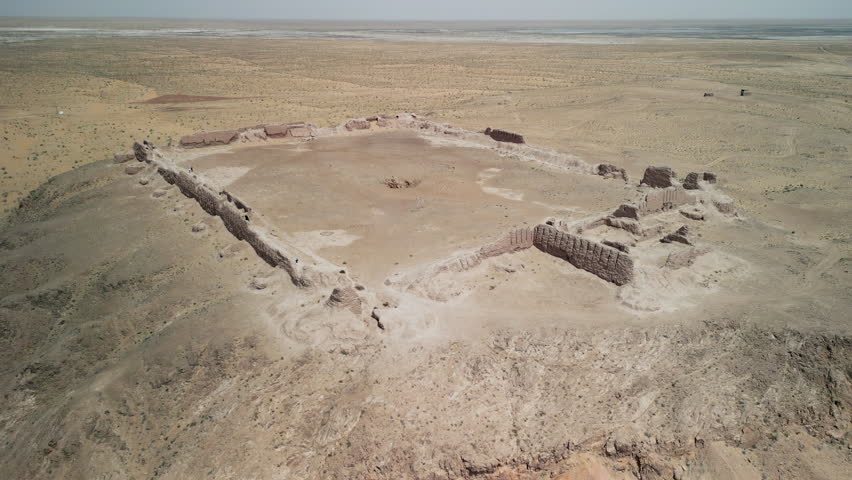 Squared ancient ruins of Ayaz-Kala seen from high above. Walls remain visible on desert plateau. Visitors walk the perimeter amid scattered sand and sparse vegetation.
