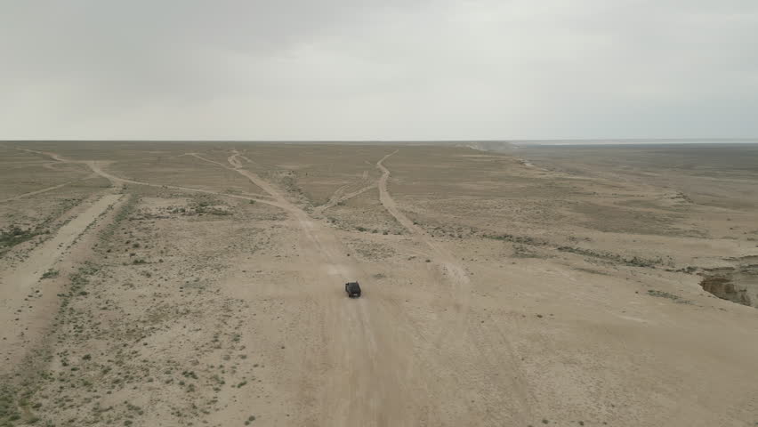Aerial view of a solitary offroad vehicle as it moves through a network of winding dirt roads. Endless arid steppe stretches beyond, with dry earth and scattered desert plants
