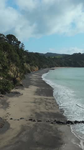 Scenic view of a dark sand and pebble beach with gentle waves and a lush green hillside under a cloudy sky. Natural coastal landscape. Orere Point, Auckland, New Zealand