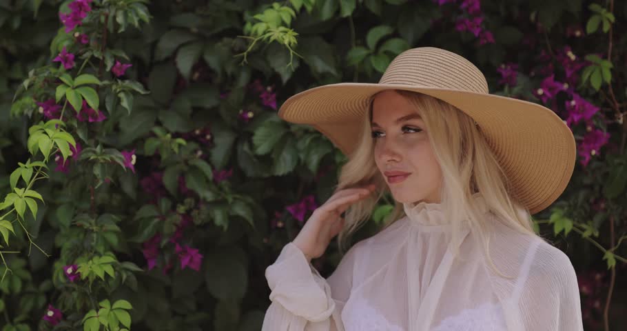 Blonde woman in sheer white summer outfit and wide-brim hat poses gently near floral garden wall. Ideal for fashion, skincare, beauty, influencer, or nature-inspired promo content.