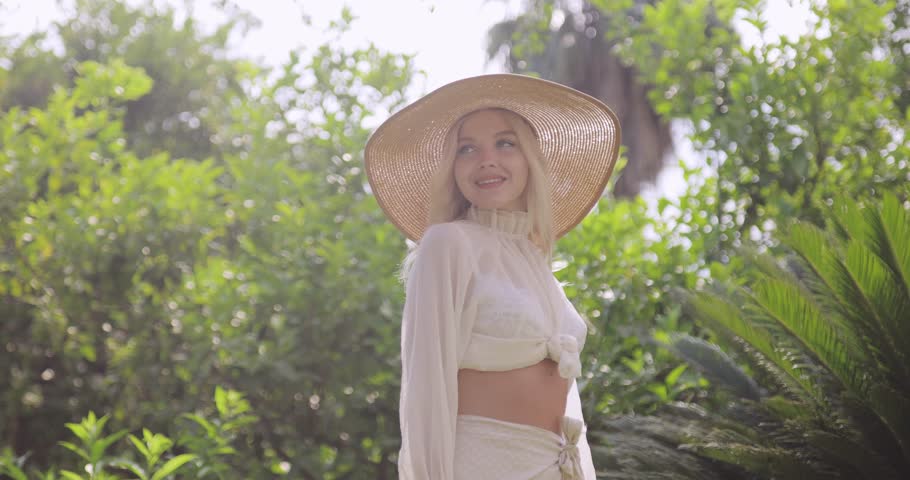 Stylish Caucasian woman in trendy summer outfit and wide-brim hat posing outdoors in tropical garden. Concept for fashion promo, vacation, lifestyle, or beauty advertisement.