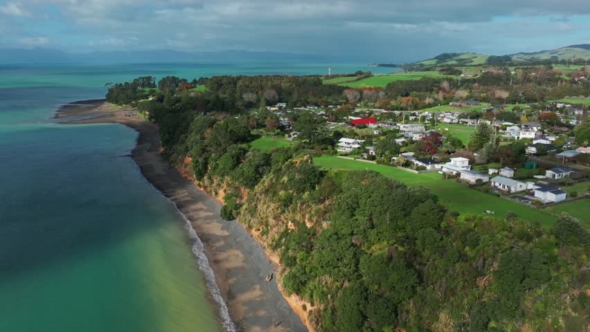 Aerial view of a coastal town and beach with turquoise water, cliffs, and green hills in Orere Point. People walk along the shore.Auckland, New Zealand
