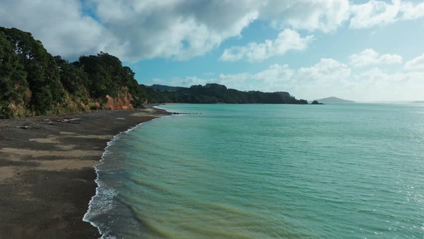Scenic view of a rugged coastline with a dark sand beach, green trees on cliffs, and turquoise sea under a cloudy sky. Orere Point, Auckland, New Zealand