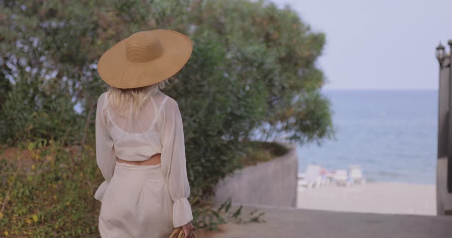 Caucasian woman in white sheer outfit and straw hat walking gracefully toward a serene beach. Ideal for travel, vacation, fashion, and lifestyle promo with elegant summer vibes.