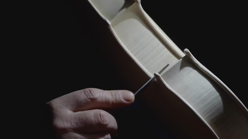 Extreme close up macro shot in slow motion of hand of master lute maker smoothing and shaping corner edges near the center bout with raw rasp to new unpainted classical violin