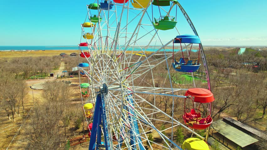 Aerial drone footage of a colorful Ferris wheel located near the shore of Kapchagay Reservoir. The bright gondolas in various colors contrast beautifully with the natural background of water