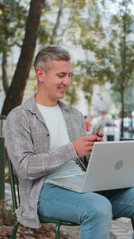 Happy rich man using laptop winner victory counting holding fan of cash money in dollar banknotes sitting on chair city street park. Young male guy tourist planning budget online shopping. Vertical