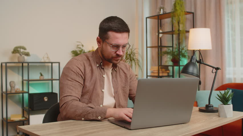 Young businessman freelancer stock trader earning holding showing bitcoins after online monitoring trading operations. Increasing wealth, financial prosperity. Guy with laptop at home office table.