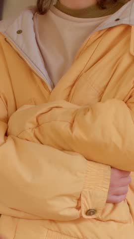 In an indoor environment, a young woman, dressed in a trendy yellow jacket, stands with her arms crossed, maintaining a neutral expression that complements her casual attire