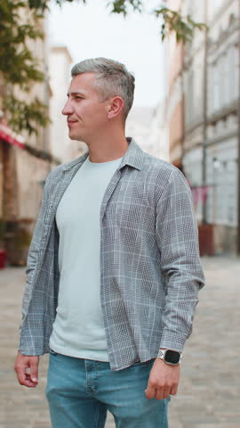 Serious dissatisfied Caucasian mature grey-haired man stands outdoors looking for someone waiting for meeting angry frustrated looks time wristwatch feeling upset about being late lateness. Vertical