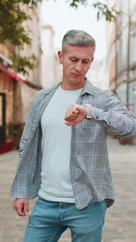Serious dissatisfied Caucasian mature grey-haired man stands outdoors looking for someone waiting for meeting angry frustrated looks time wristwatch feeling upset about being late lateness. Vertical