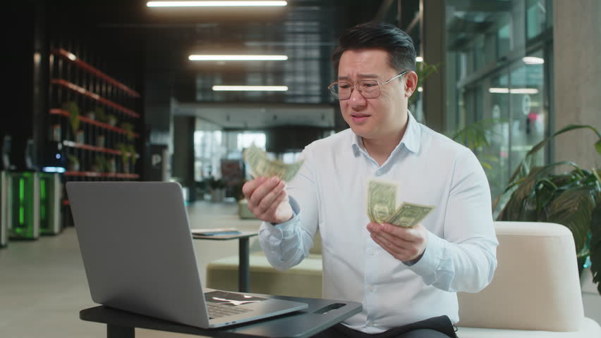 Depressed Asian businessman feeling worried about financial problem. Stressed man in shirt looking frustrated thinking of money debt, bad day, budget loss, bankruptcy sitting in modern office lobby.