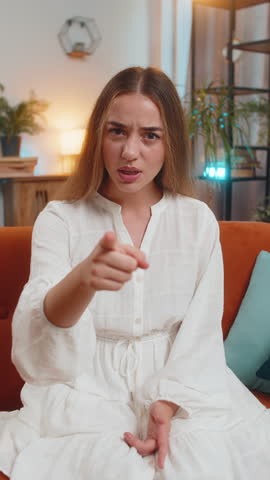 Furious young Caucasian woman sitting on sofa couch at home, angrily pointing at you through the camera, displeased and fuming. Adult Girl in serious blame mode, upset, and ready to explode. Vertical.