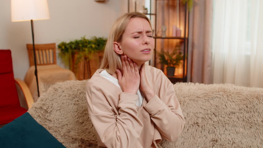 Young adult woman touches her throat on home sofa, clearly showing pain while trying to swallow. Blonde girl reacts with visible discomfort, expressing illness or inflammation in her throat area.