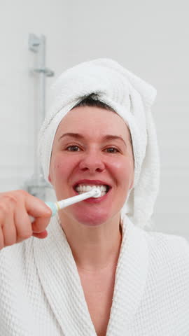 Middle-aged mature woman happily brushing teeth carefully with electric toothbrush in front of bathroom mirror. Lady smiling cheerfully, enjoying fresh daily oral care hygiene routine at home