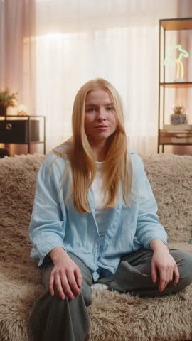 Woman on sofa reveals doubt, displaying lack of answers with a hesitant face under soft sunset light. Girl lifts shoulders at home, opening arms in unsure gesture, leaning head to side, no answer idea