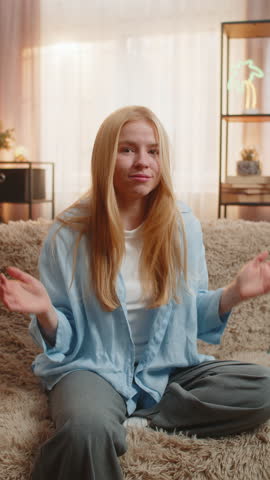 Woman on sofa reveals doubt, displaying lack of answers with a hesitant face under soft sunset light. Girl lifts shoulders at home, opening arms in unsure gesture, leaning head to side, no answer idea