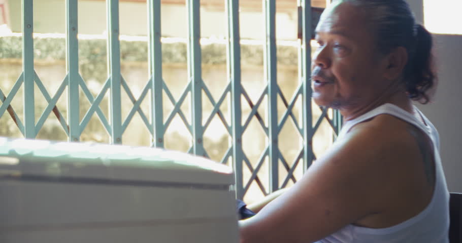 Asian man which is senior, sits beside a washing machine behind his house. Surrounded by concrete and daily items, he does his laundry in silence, his face lost in memory and quiet thought.