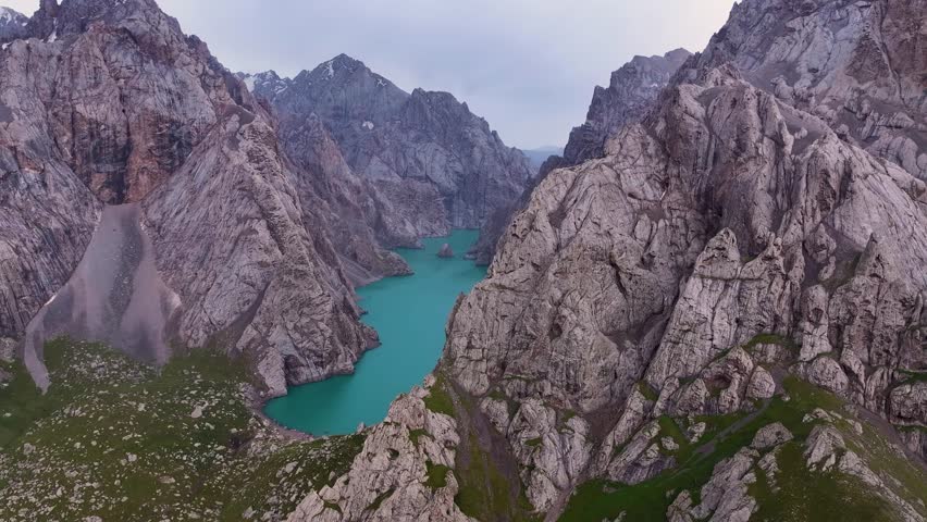 Kel Suu Lake, with its striking turquoise waters, is nestled deep within a dramatic landscape of towering, rugged rocky mountains under a cloudy sky