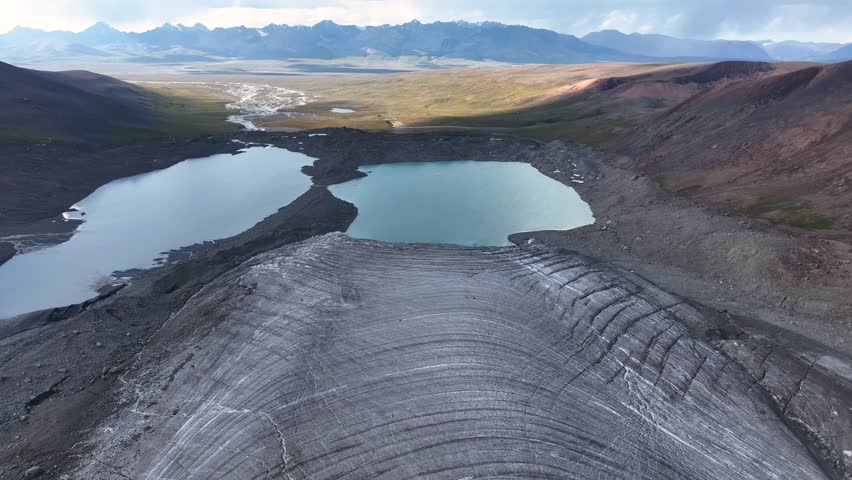 An aerial view captures a vast glacier, two serene glacial lakes, and a wide mountain valley with a river in the distance