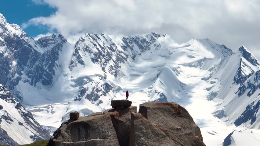 A lone person stands on a large rock, gazing at the breathtaking snow-covered mountains under a bright sky. The camera is orbiting
