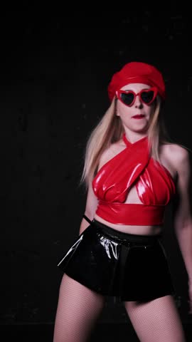 Beautiful sexy girl in red latex top, black latex skirt and red beret with red heart-shaped glasses. Posing and dancing on dark background
