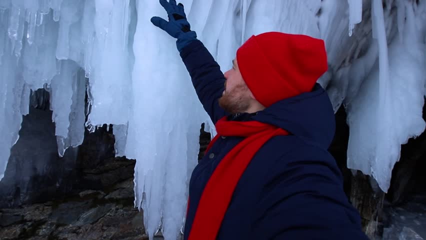 Adventure travel in winter, tourist man with red scarf makes selfie background ice grotto and cave, frozen icicles of Lake Baikal.