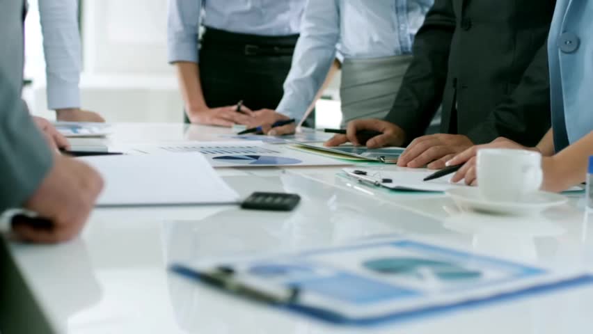Dynamic footage showing a group of business professionals gathered around a desk during a corporate strategy meeting. The team is pointing at charts, analyzing documents, and actively collaborating. 