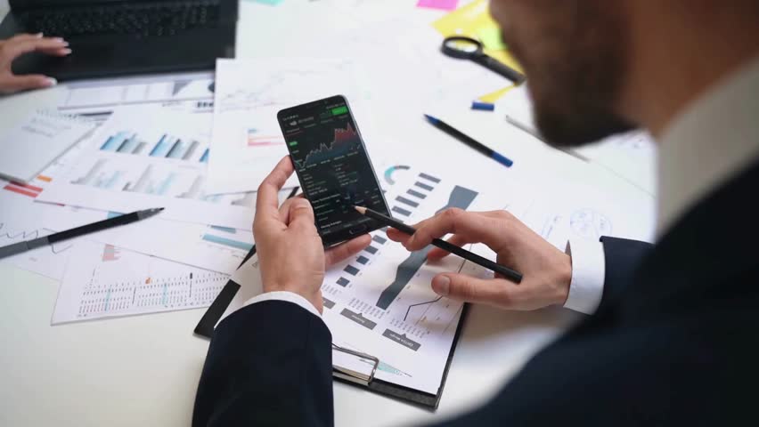 A financial analyst uses a smartphone to analyze stocks amid graphic reports.