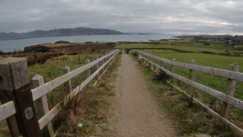 Coastal trail to Great Pollet Sea Arch in Donegal, Ireland. Gravel path with wooden fence, wild cliffs, and Atlantic Ocean under a dramatic sky.