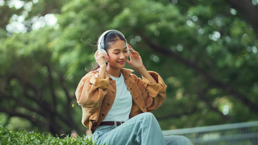 Happy beautiful young asian woman relaxing and enjoying music with headphones in the park – peaceful outdoor moment with gentle breeze, smiling female feeling calm and joyful in nature