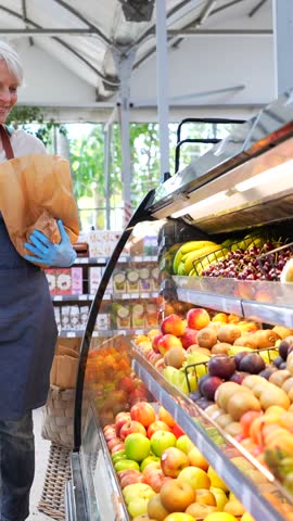 Experienced senior greengrocer carefully filling paper bag with ripe, colorful produce while working at vibrant local fruit market stall