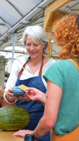 Senior greengrocer carefully selecting ripe melon, engaging with customer paying through modern contactless credit card technology at bustling local market stall