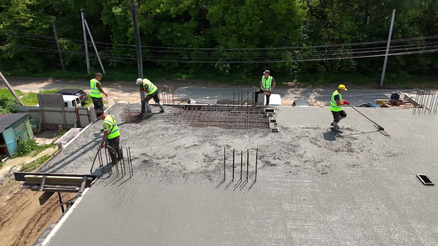 Construction workers pouring and leveling concrete slab. Aerial view of construction crew wearing safety vests as they pour, spread, and level wet concrete on a building roof.