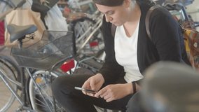 Young woman using her smartphone while sitting beside her bicycle, likely waiting for someone or something in an urban environment, embracing a casual lifestyle slow motion shot - Powered by Shutterstock - Get 15% off with code: PIKWIZARD15