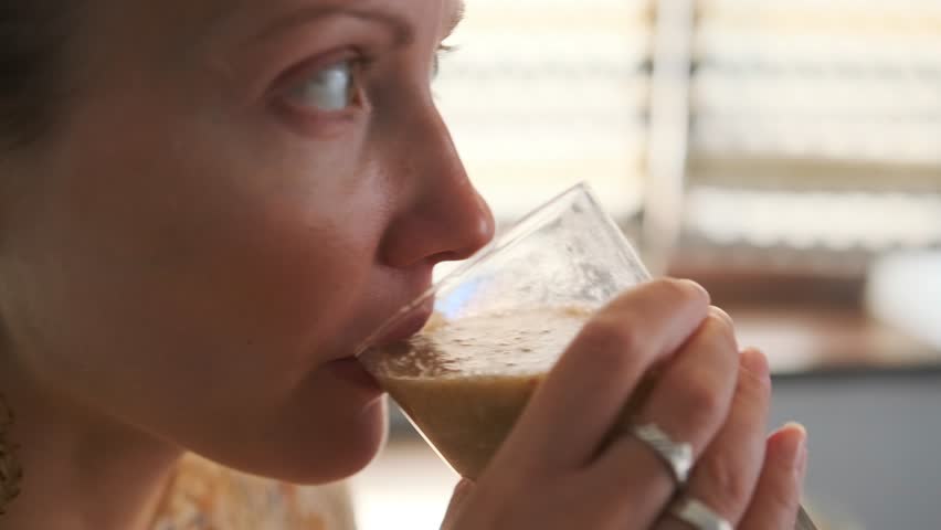 Close-up side view of a woman drinking a healthy smoothie from a glass in a bright home interior. Peaceful moment of wellness and self-care in the morning light.