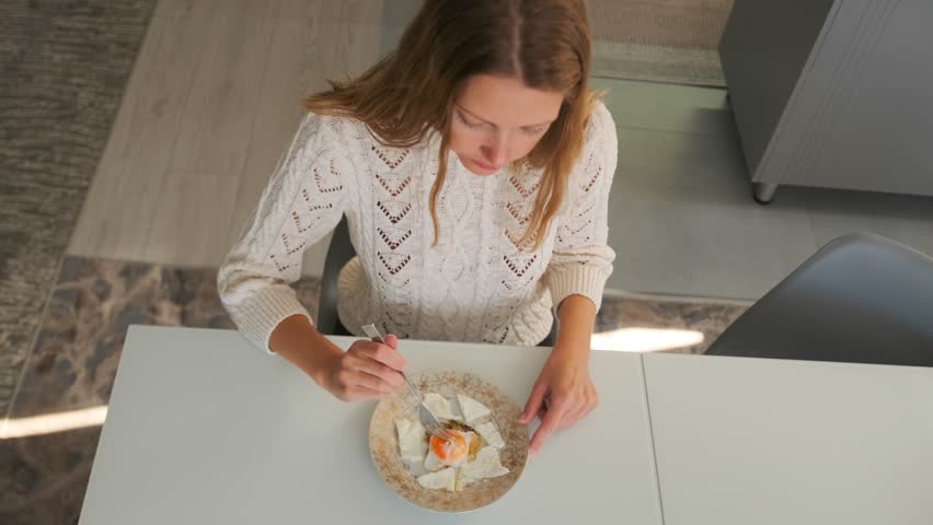 Top view of a young woman eating a perfectly fried egg with a fork while sitting at a table in a bright and cozy room. Enjoying a home-cooked meal in the morning light.