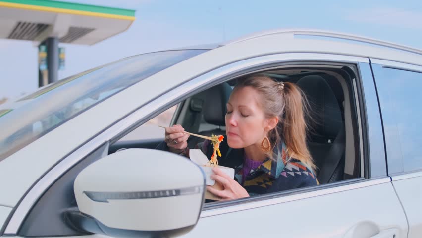 Young woman eats noodles with chopsticks in her car while parked at a gas station, quick stop for food on a busy day. Realistic and relatable moment of modern travel life.
